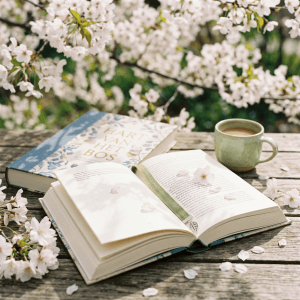 Open book with flower bookmark, closed book, coffee cup, and cherry blossom petals on rustic wooden table
